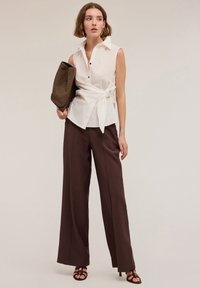 Blouse blanche sans manches avec une taille nouée, comportant un col et des boutons noirs, associée à un pantalon large taille haute marron.