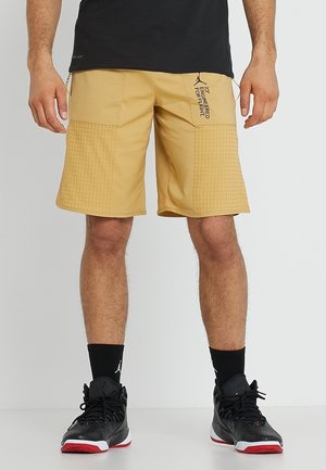 Beige sportieve shorts met een textuurpatroon, ritszakken en bedrukte accenten. G gedragen met zwarte sneakers en sokken.