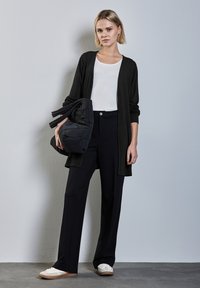 Musta pitkä neuletakki valkoisen t-paidan päällä, yhdistettynä korkeavyötäröisiin mustiin housuihin. Hän pitää tummasta kirjotusta laukusta. Kengät ovat valkoiset, jossa on beige pohja.