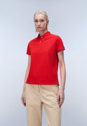 Young person with short blonde hair wearing a red short-sleeve polo shirt and beige pants against a plain light background.