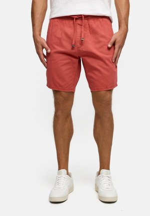 Mann trägt lachsfarbene Shorts mit Kordelzug, weiße Turnschuhe und ein weißes T-Shirt, steht vor einem einfarbigen hellen Hintergrund.