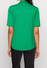 Personne portant une chemise collée verte à manches courtes et un pantalon noir, vue de dos contre un fond uni.