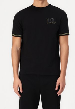 Schwarzes Baumwollt-Shirt mit kurzen Ärmeln, das gelbe Streifenakzente an den Ärmeln und ein grafisches Logo auf der Brust aufweist. Glatte Textur.