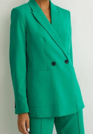 Femme portant un blazer croisé vert vif avec des boutons noirs et un pantalon vert assorti sur un fond clair uni.