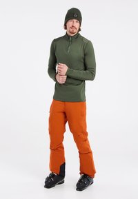 Herren Langarm-Basislayer in Olivgrün mit Viertel-Reißverschluss, kombiniert mit leuchtend orangefarbenen Schneehosen, die schwarze Akzente und Skischuhe aufweisen.