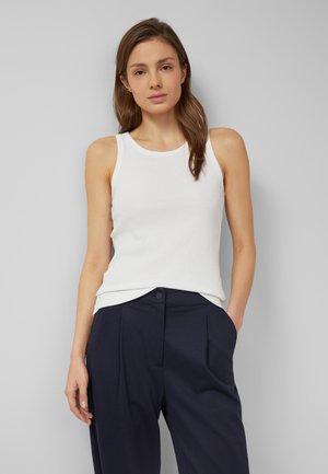 Frau mit langen braunen Haaren, die ein weißes ärmelloses Oberteil und dunkelblaue Hose trägt, stehend vor einem schlichten hellgrauen Hintergrund.