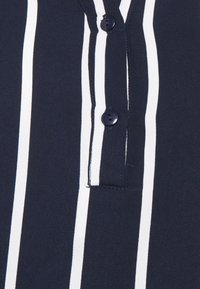 Tkanina s námořnickými modrými a bílými pruhy, se dvěma tmavými knoflíky, s klasickým límcem a kusem látky poblíž légou knoflíku.