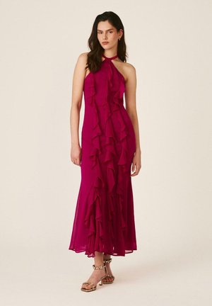 Mujer de pie con un vestido magenta sin mangas, largo hasta el suelo, con volantes en cascada y sandalias doradas de tacón alto, sobre un fondo liso.