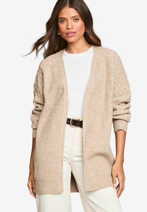 Beige cardigan with ribbed texture and patterned sleeves, open front, soft material, worn over a white top and paired with cream pants.