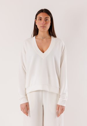 Donna con capelli lunghi e scuri che indossa un maglione bianco a V ampio e pantaloni abbinati a gamba larga, in piedi davanti a uno sfondo chiaro.