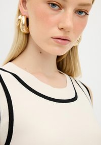 Top aderente senza maniche in tessuto a maglia color crema, con bordi neri e scollatura arrotondata. La modella indossa grandi orecchini a cerchio in oro.