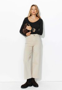 Haut à manches longues noir avec détail en dentelle, pantalon large beige, et bottines brunes. Tenant un sac en léopard brun.