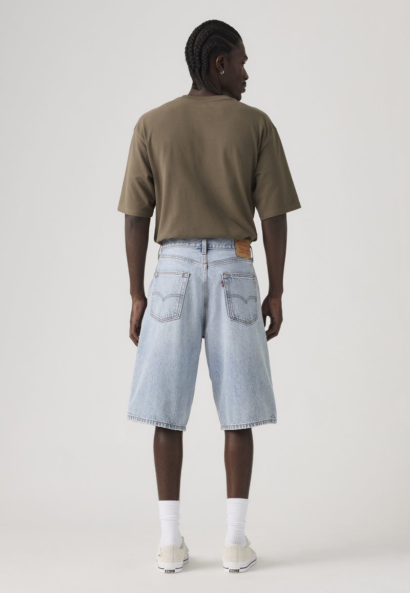 Mann mit geflochtenem Haar steht mit dem Rücken zur Kamera, trägt ein olivgrünes, übergroßes T-Shirt, hellblaue Jeansshorts, weiße Socken und Turnschuhe.