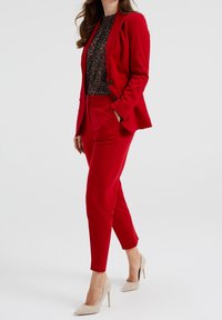 Czerwony dopasowany garnitur z taliowanym żakietem i zwężanymi spodniami. Teksturowana czarna bluzka pod spód. Modelka nosi beżowe szpilki. Gładki materiał, ostre linie.