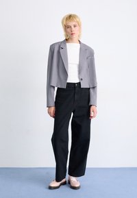 Osoba stojaca na modrej podlahe, oblečená v skrátenom šedom blejzri, bielom tričku, čiernych širokých nohaviciach a svetloružových topánkach pred bielou stenou.