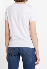 T-shirt blanche à manches courtes, avec un col rond rose. L'arrière-plan montre un jean en denim bleu avec des poches arrière.