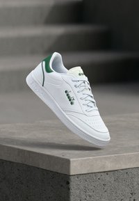 Αθλητικό παπούτσι από λευκό δέρμα με πράσινες λεπτομέρειες στη φτέρνα και τη γλώσσα. Διαθέτει διάτρητο μπροστινό μέρος, κορδόνια και υφασμένο λαστιχένιο πέλμα.