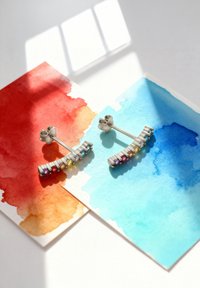 Silberne Ohrringe mit mehrfarbigen Edelsteinen, die in einer geschwungenen Linie angeordnet sind, dargestellt auf roten und blauen Aquarellhintergründen.