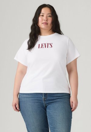 Frau mit langen dunklen Haaren, die ein weißes Levi's-T-Shirt und blaue Jeans trägt, steht vor einem schlichten hellen Hintergrund.