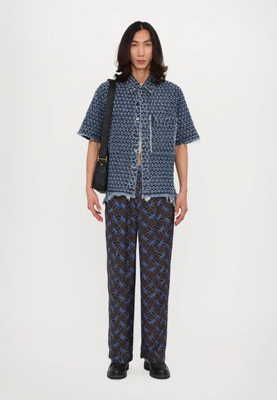 Blue patterned short-sleeve shirt with frayed edges, paired with brown trousers featuring a blue geometric pattern. Black bag and shoes.