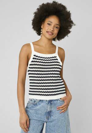 Femme avec des cheveux afro bouclés portant un haut sans manches rayé noir et blanc et un jean en denim bleu clair, debout avec une main dans la poche.