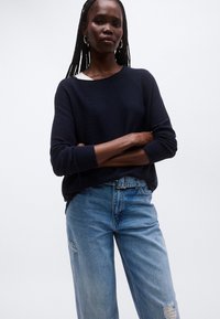 Merensininen neulepusero, jossa on resorit hihansuissa, puettuna valkoisen topin päälle ja yhdistetty vaaleansinisiin revittyihin farkkuihin. Neuleessa on väljä istuvuus.