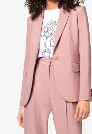 Nainen pukeutuneena pölyisen vaaleanpunaiseen mittatilausbleiseriin ja siihen sopiviin korkeavyötäröisiin housuihin, valkoisen paidan päällä, jossa musta kukkainen viivapiirroskuvio.