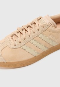 Tan suède sportieve schoen met drie beige strepen, textuur bovenwerk en rubberen zool. Bevat een reliëflogo op de tong en gestikte accenten.