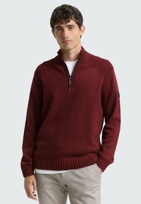 Dunkelroter Pullover mit Reißverschluss am Kragen, Rippb�ündchen an Ärmelabschlüssen und Saum, gefertigt aus strukturiertem Stoff. Kombiniert mit einem weißen Hemd darunter.