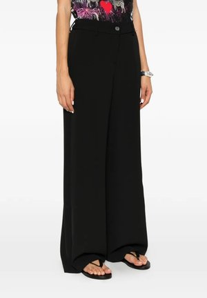 Donna che indossa pantaloni neri larghi e morbidi e infradito nere, in piedi con le mani rilassate ai lati su sfondo bianco.