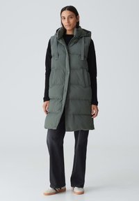 Grüne, puffy Weste mit Kapuze, mit Knopfleiste, Seitentaschen und verstellbaren Schulterriemen, getragen über einem schwarzen Pullover und dunklen Hosen.