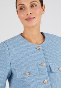 Blauwe textuurjas met ronde hals, voorzien van gouden knopen en twee voorzakken. De stof heeft een geweven patroon en een subtiele glans.