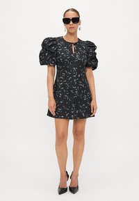 Čierne šaty s kvetinovým vzorom, krátkymi puff rukávmi, a výstrihom v tvare kľúča, vyrobené z textúrovaného materiálu. Nosené s čiernymi slnečnými okuliarmi a ihlovými topánkami.
