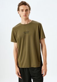 Camiseta deportiva de manga corta color verde oliva, hecha de material suave y ligero. Presenta un cuello redondo y texto impreso en negro: "NO TE DETENGAS HASTA QUE ESTÉS ORGULLOSO."