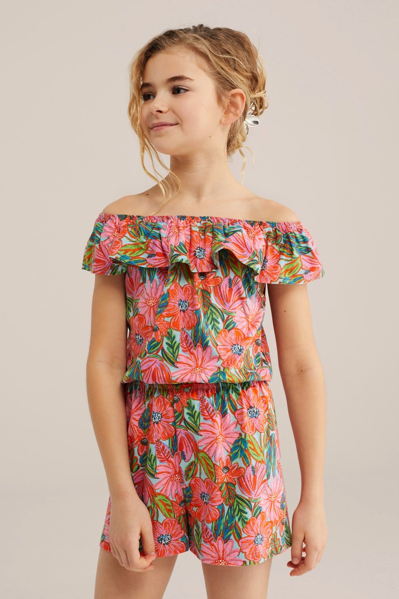 Niña joven con blusa floral colorida de hombros descubiertos y shorts a juego, cabello recogido de forma suelta con una pinza, sobre fondo liso.