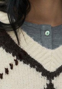 Maglione lavorato a maglia con una base crema e motivi geometrici marroni scuri, design a scollo a V, e un colletto a coste grigio con un bottone azzurro chiaro.