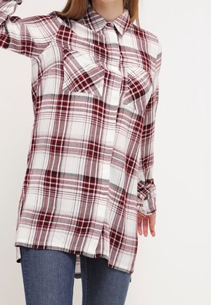 Femme portant une chemise à manches longues, rouge et blanche à carreaux, boutonnée, avec deux poches poitrine, associée à un jean bleu.