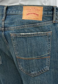 Jean en denim bleu avec une finition texturée, présentant une étiquette en cuir à la ceinture et des surpiqûres orange le long des poches et des coutures.