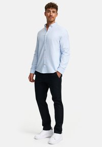 Uomo in piedi con le mani in tasca, che indossa una camicia azzurra chiara con bottoni, pantaloni neri e scarpe da ginnastica bianche su uno sfondo semplice.