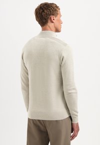 Man die met zijn rug naar de camera staat, gekleed in een lichtbeige gebreide trui met geribde kraag en manchetten, gecombineerd met een paarse broek tegen een effen witte achtergrond.