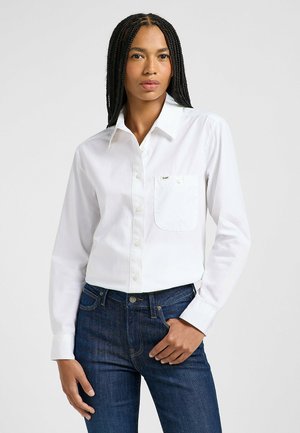 Chemise blanche à boutons en tissu coton, avec une poche avant et des manches longues, associée à un jean en denim bleu foncé.