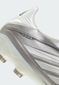 Gros plan sur une chaussure de football Adidas Copa argentée et blanche, mettant en avant la tige texturée, les trois bandes et la semelle à crampons.