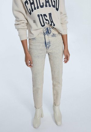 Personne portant un jean taille haute beige à effet délavé acide, un sweat-shirt crème "Chicago USA" et des bottines crème à talons hauts sur un fond uni.