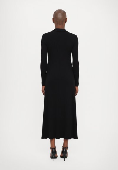 Melns, ribots garas piedurknes kleita ar augstu apkakli, pieguļošs priekškars un plūstošs svārku garums līdz potītēm; kombinēta ar melniem augstpapēžu apaviem.