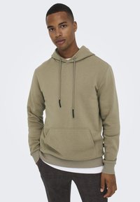 Olive green hoodie made of soft fabric, featuring a drawstring hood, kangaroo pocket, and ribbed cuffs. Worn over a white shirt.