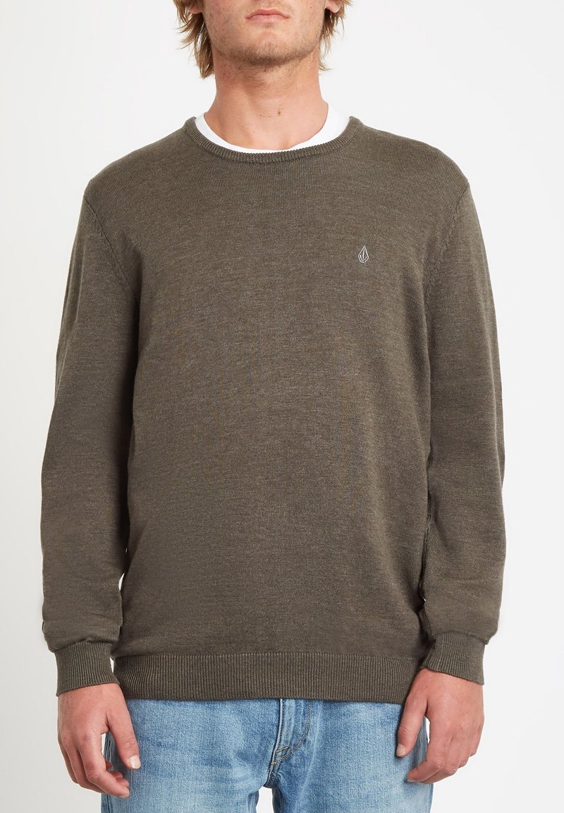 Homme portant un pull en tricot vert olive par-dessus un tee-shirt blanc avec un jean bleu clair, debout devant un fond uni.