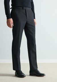 Schwarze, maßgeschneiderte Hose mit slim fit, ausgestattet mit flachem Frontbereich, Bügelfalten und glattem Stofffinish, getragen mit schwarzen Loafers.