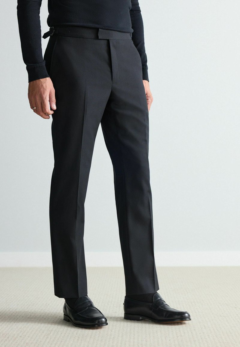 Schwarze, maßgeschneiderte Hose mit slim fit, ausgestattet mit flachem Frontbereich, Bügelfalten und glattem Stofffinish, getragen mit schwarzen Loafers.