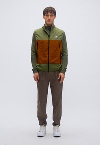 Πράσινο και καφέ γιλέκο με φερμουάρ και ψηλό γιακά, με λογότυπο. Συνδυάζεται με μπεζ παντελόνι και λευκά αθλητικά παπούτσια, σε απλό φόντο.