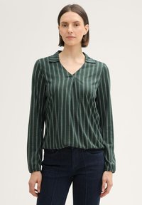 Grüne Bluse mit langen Ärmeln und V-Ausschnitt, mit vertikalen weißen Streifen und einer weichen Textur. Kombiniert mit dunklen Jeans.
